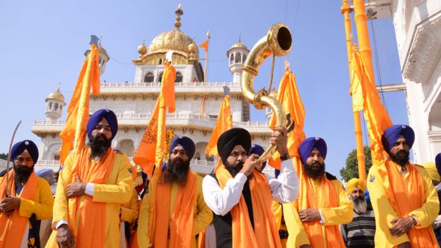 An Indian Sikh devotee plays a musical instrument as he walks with Punj Pyara holding flags